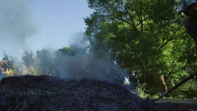 一个四十多岁的高加索人将树叶，木棍和树枝和灰烬耙到户外的一个大燃烧燃烧堆上
