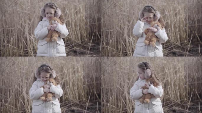 走失的白人小女孩和泰迪熊在户外哭泣的画像。悲伤孤独的孩子站在灰色的灌木丛后面拥抱着玩具。问题，沮丧，