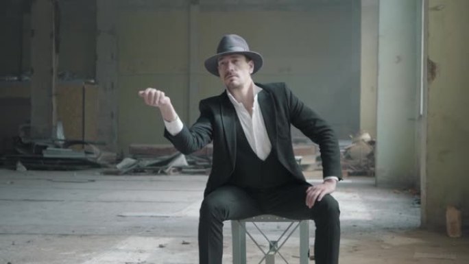 一个男人坐在被毁的车库的椅子上戴着软呢帽，穿着古典西装，抛硬币。