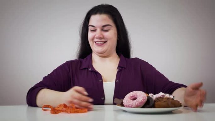 微笑的高加索肥胖女孩坐在糖果和卷尺前的肖像。胖女人把卷尺移到一边，拿盘子和甜点。肥胖，节食，自我控制