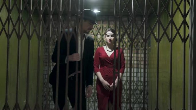穿着西装和帽子的高个子男人打开门，在黑暗的房间里经过一个穿着优雅红色连衣裙的自信漂亮的女孩。