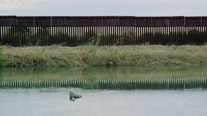 日落时分，在墨西哥和美国之间的部分阴天，垃圾漂浮在一条与钢板条边界墙平行的运河上，上面是剃刀线 (在