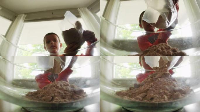 一个可爱的6岁白人男孩白天在室内厨房烘焙时，把巧克力蛋糕粉从袋子里倒进一个大的玻璃碗里