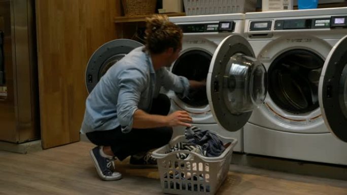 英俊的年轻人从烘干机里拿出衣服，把它们放在洗衣篮里，在自助洗衣店微笑着