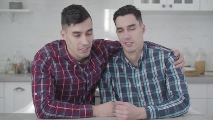 两个成年高加索双胞胎兄弟的特写镜头互相交谈，转向镜头并微笑。积极的年轻人在家里摆姿势。友谊，家庭，同