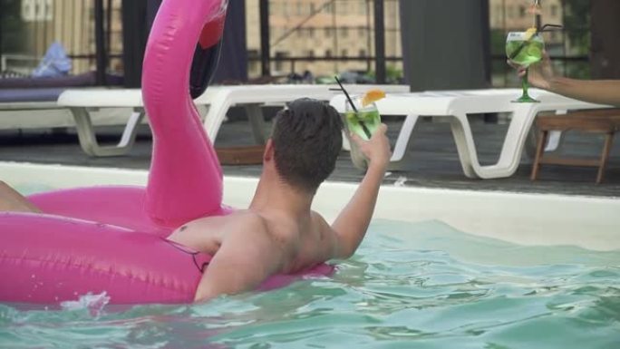 年轻有趣的男人在充气火烈鸟边游泳，在游泳池里热黑发和可爱的性感金发女郎