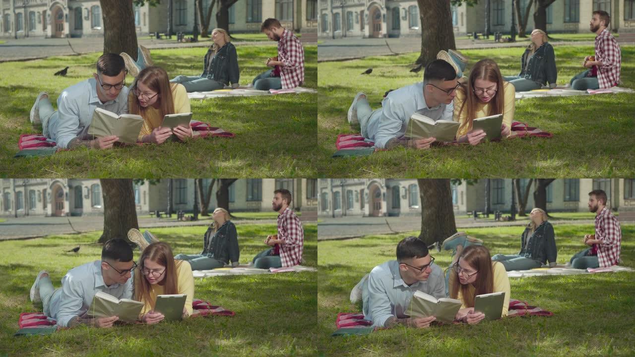 聪明的年轻男孩和女孩躺在绿色草坪上的眼镜上，读书，作为相爱的夫妇在后台玩夏威夷四弦琴。被吸收的高加索