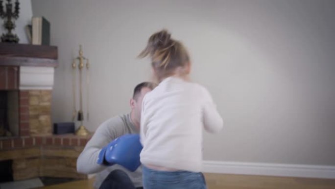 镜头跟着黑发小高加索女孩走到年轻人面前，把脸藏在拳击手套后面，给他拳击。开朗的女儿在家和可爱的父亲玩