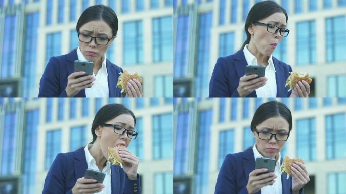 年轻的女经理匆忙吃不健康的汉堡，在电话上查看新闻