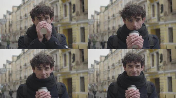 正面英俊的高加索男孩喝咖啡，对着镜头眨眼的特写肖像。微笑的年轻人站在秋天的城市街道上。休闲，生活方式