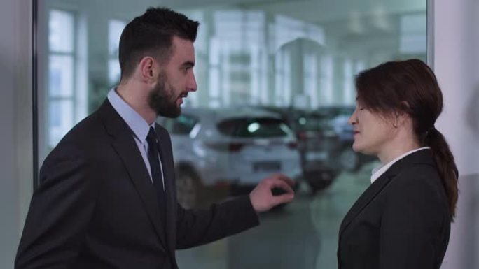 严肃的高加索男人和女人站在汽车经销店聊天的侧视图。男性经销商向女性客户解释汽车规格。电影院4k Pr