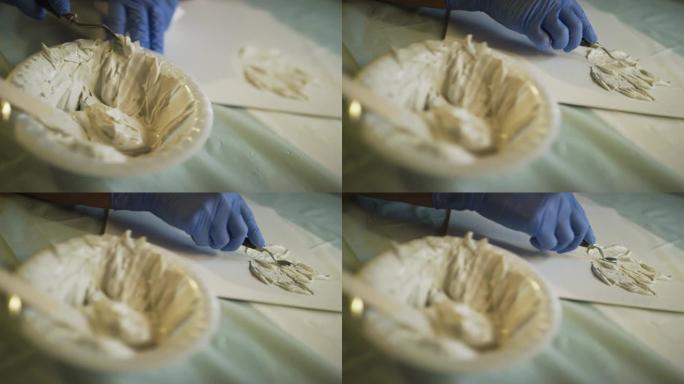 一位戴着防护手套的艺术家的机架焦点镜头将调色板刀滴入一个湿的干墙泥/腻子的聚苯乙烯碗中，并用调色板刀