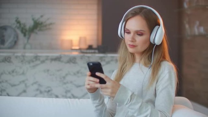 年轻的女孩在看智能手机的屏幕的耳机听音乐。在家放松