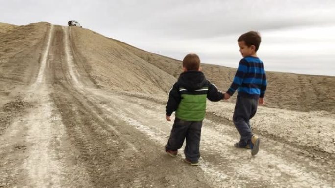 秋天，两个高加索兄弟 (两岁和三岁) 在阴暗的天空下走在科罗拉多州高高的沙漠中的一条土路上，手牵手