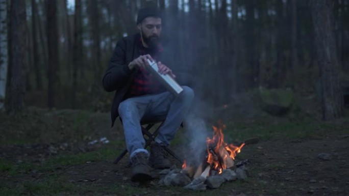 大胡子的人坐在篝火附近的森林里，把饮料放在热水瓶里的杯子里。野蛮的未刮胡子的家伙在森林里的火旁休息。