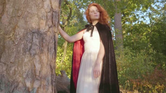 美丽的白人女孩，红头发，穿着白色长裙，黑色长袍，红色衬里，站在树旁边