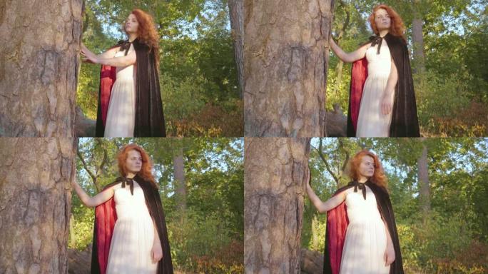 美丽的白人女孩，红头发，穿着白色长裙，黑色长袍，红色衬里，站在树旁边