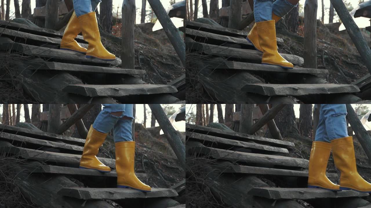 穿着黄色橡胶靴和蓝色牛仔裤的人腿在户外的木制楼梯上走下。穿着橡胶靴的脚从森林的木制楼梯上下来。
