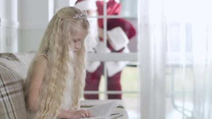 侧视图，漂亮的白人金发女孩坐在沙发上看书，圣诞老人来到玻璃入口门口，向内看，敲门。儿童合上书和挥手。