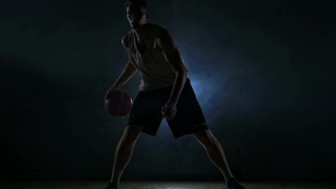 盘带篮球运动员在球场上，球在黑暗的房间里，背光在烟雾中慢动作