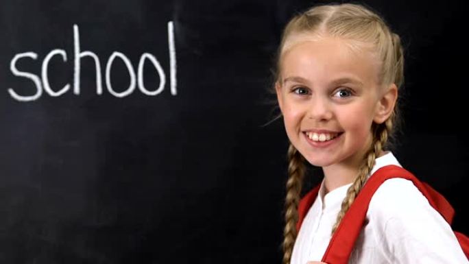 背着背包的小女孩对着镜头微笑，学校单词写在黑板上