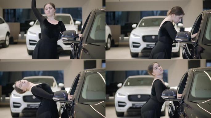 迷人的高加索芭蕾舞演员弯下腰在黑色汽车旁边。在汽车经销店跳芭蕾舞的年轻漂亮女人。汽车陈列室里的苗条芭