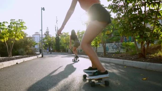 滑板上的两个女性朋友在度假胜地下坡骑行，开心地移动和微笑。沿着棕榈树的道路