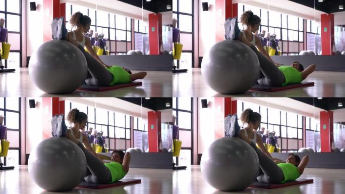 美丽的私人教练帮助成熟的女人，同时在垫子上锻炼她的腹肌，并将腿靠在球上