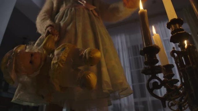 白人女孩穿着礼服站在烛台旁边，拿着蜡烛和洋娃娃的仰视图。晚上独自在烛光下的怪异孩子。恐惧，恐惧，奇怪