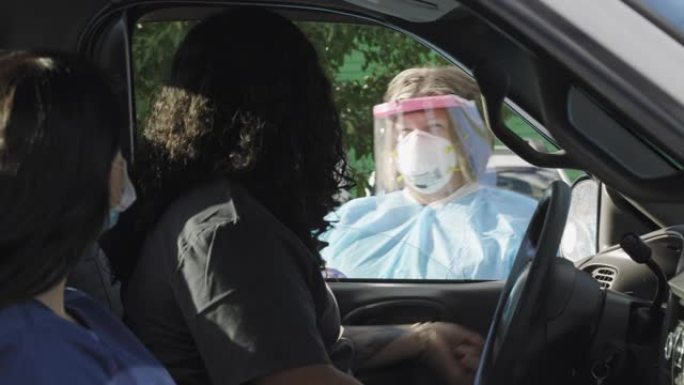 一名身穿长袍、戴着手术面罩、手套和面罩的女护士走上卡车，与一名身穿卡车的男性和一名女性在开车新型冠状
