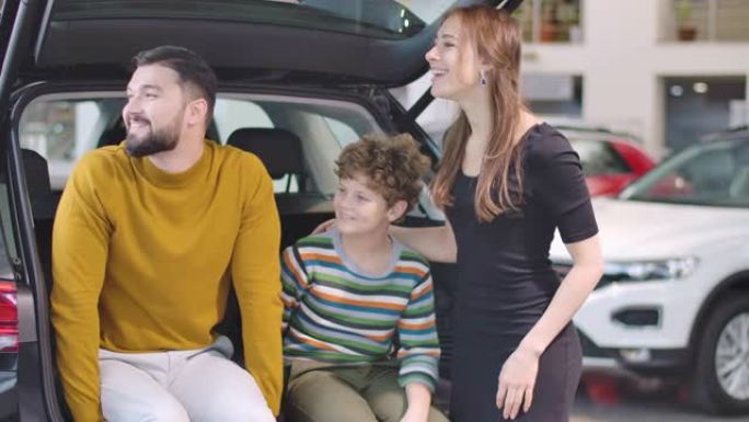 年轻的大胡子高加索人与儿子和妻子坐在敞开的汽车后备箱中，望着远处。欢快的家庭在展厅租车或买车。汽车租