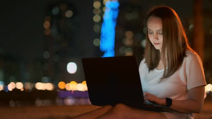 年轻的女孩设计师晚上在棕榈树下在外面的笔记本电脑上远程工作