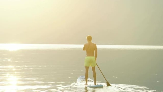 年轻人在平静的湖上朝着日出站起来桨板