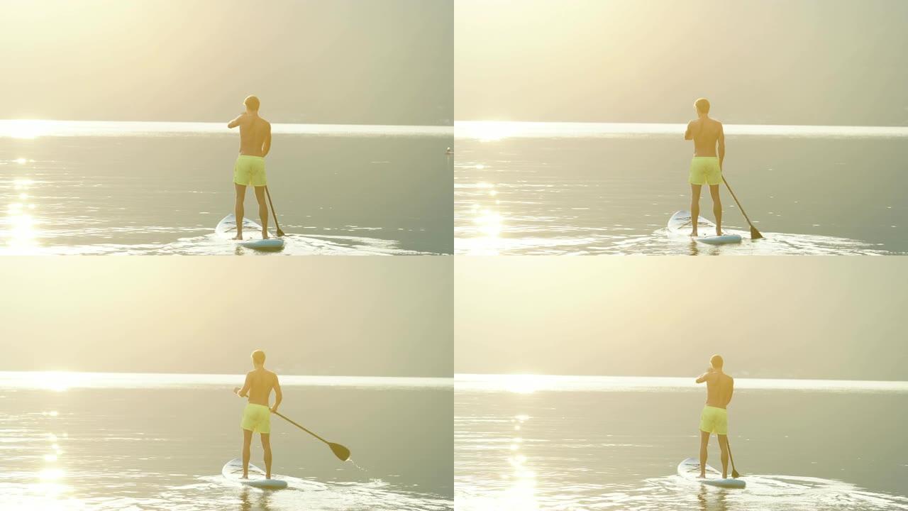 年轻人在平静的湖上朝着日出站起来桨板