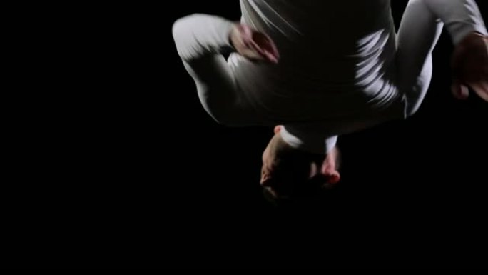 穿着白色衣服的黑色背景的男性体操运动员从下方飞入框架，在空中旋转并以慢动作翻转。比赛中的体操运动员。