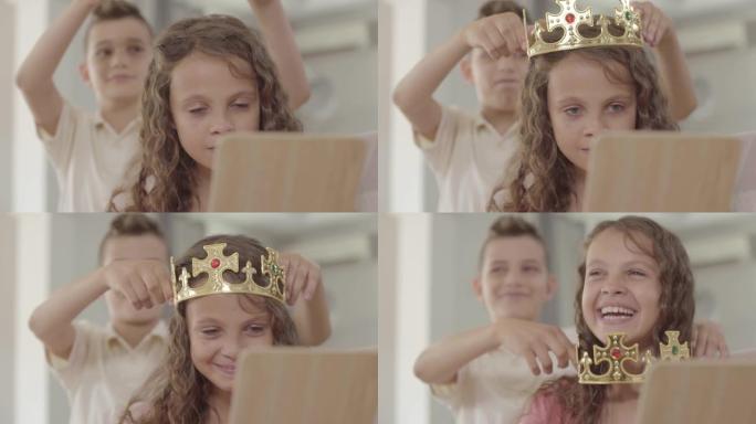可爱的男孩把皇冠戴在看着镜子的可爱的卷发女孩的头上。