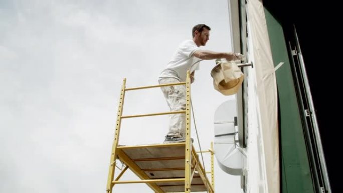一位三十多岁的高加索专业画家在部分多云的天空下站在脚手架上时，使用喷漆器对金属仓库的外部进行油漆