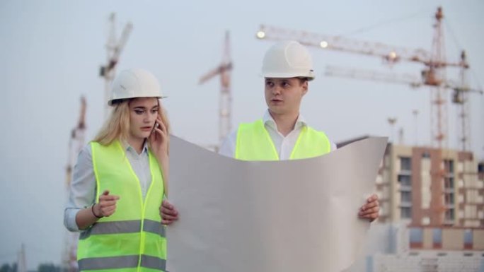 两名建筑工人戴着头盔和背心站在正在建造的建筑物的背景上，一名妇女与客户通电话
