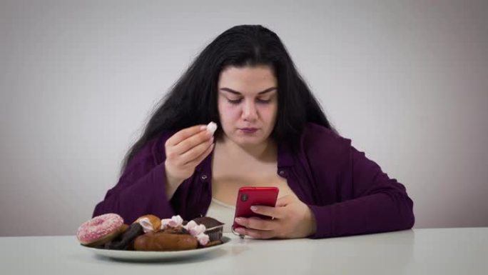 使用智能手机和吃甜食的集中高加索肥胖女孩的肖像。胖年轻女子有超重问题。不健康的节食，生活方式，肥胖。