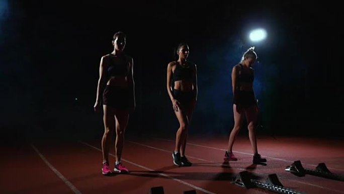三个穿着黑色衣服的女孩在起跑板上，在灯光下开始比赛，并朝着终点奔跑