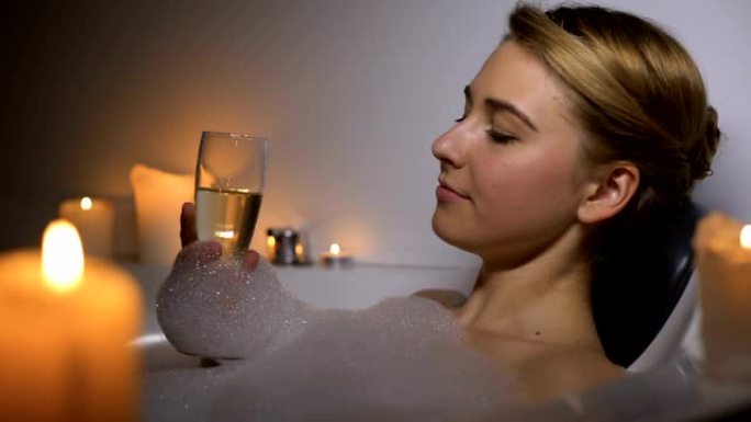 梦幻般的女人躺在泡沫泡沫和蜡烛的浴缸里，喝香槟