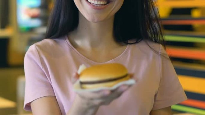 微笑的女人在镜头前展示美味可口的汉堡，有肥胖的风险