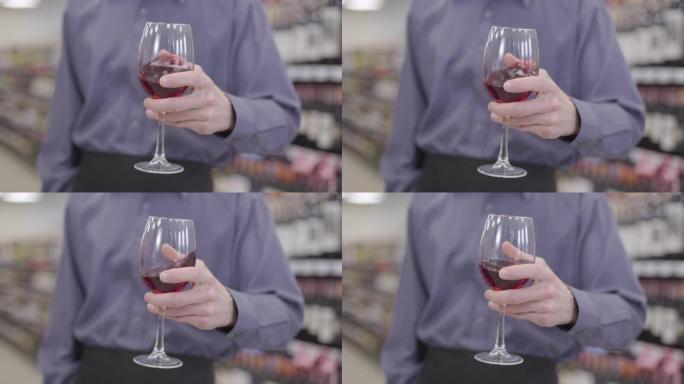 男性高加索人手摇酒杯的特写镜头，里面有红酒。无法辨认的侍酒师在酒精商店里喝昂贵的饮料。控制，品尝，酿