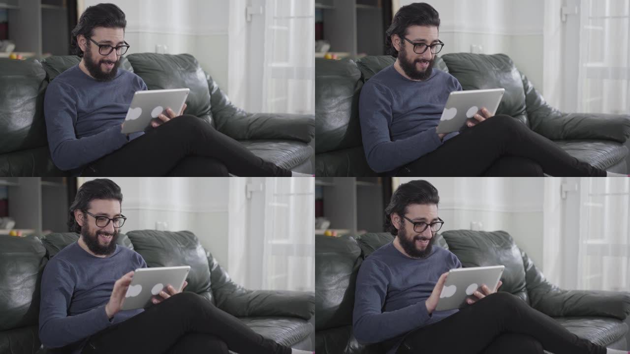 年轻的白人男子，黑发和胡须戴着眼镜，用平板电脑坐在沙发上。使用社交媒体的聪明人。网络成瘾，脸书，in