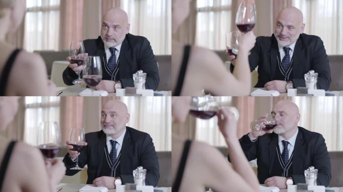 高级秃头大胡子男人与穿着黑色连衣裙的金发优雅女人碰杯，喝着红酒。幸福英俊的丈夫在豪华餐厅与妻子约会的
