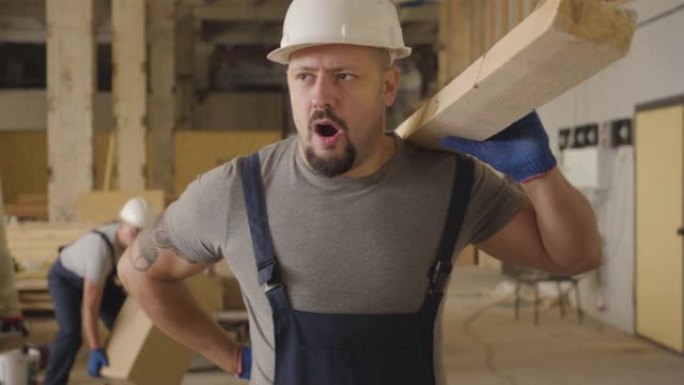 身穿白色防护头盔的肌肉发达的高加索人拿着木板在建筑工地行走。建筑商因为背部疼痛而停下来，他的同事跑来