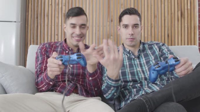 两个积极的白人男子参加电子游戏。穿着红色衬衫的家伙在放松和大笑时向他的双胞胎兄弟伸出手指。男人周末在