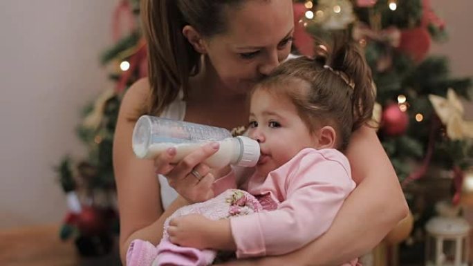 圣诞节那天，母亲在圣诞树下的瓶子里用牛奶喂养她可爱的女婴
