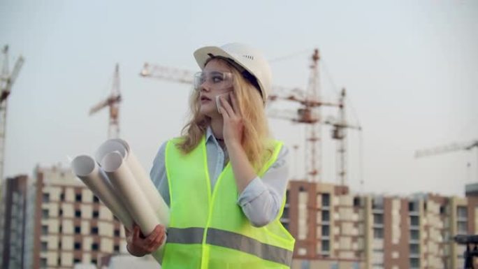 女工程师设计师与承包商通电话，手里拿着图纸，背景是在建建筑物和起重机。
