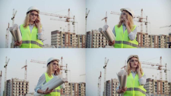 女工程师设计师与承包商通电话，手里拿着图纸，背景是在建建筑物和起重机。